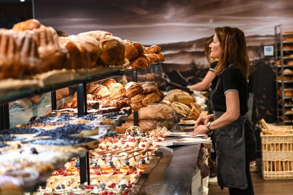 Pain chaud et pâtisserie dans une boulangerie, femme sélectionnant une tarte, viennoiseries et desserts sucrés, ambiance chaleureuse, gourmandise assurée, La Bresse.